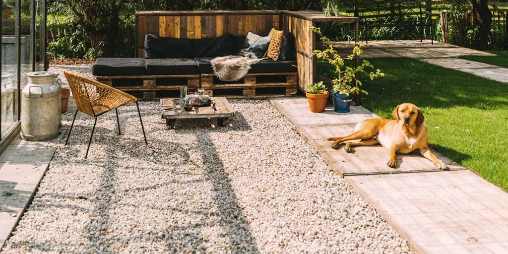 This is a picture of a dog sitting outside by a patio made out of gravel and rocks.